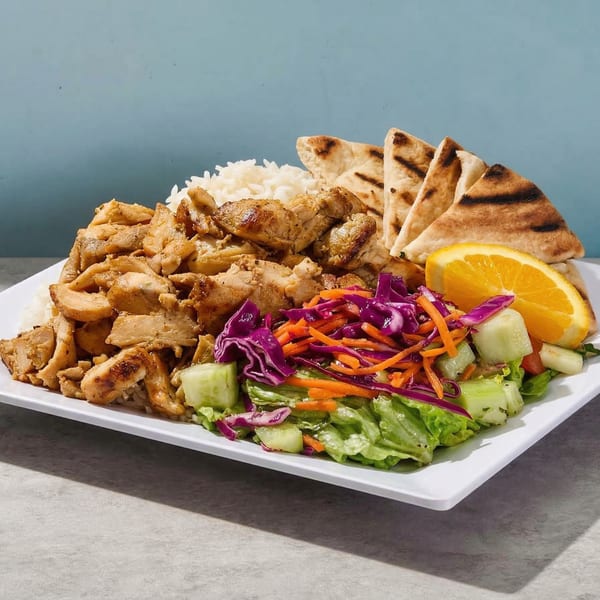 A white plate holds grilled chicken, white rice, salad with red cabbage, pita bread, and an orange slice.